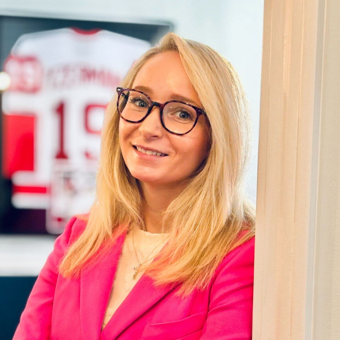 Smiling woman in glasses and pink blazer indoors.