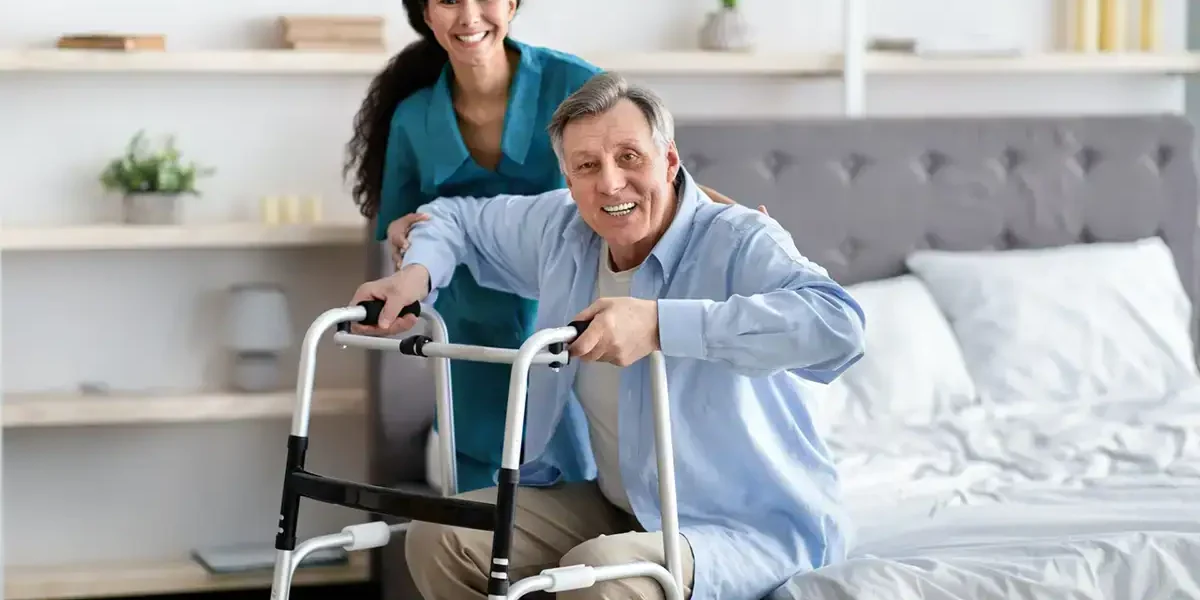 Elderly man using walker with caregiver support in a home setting.
